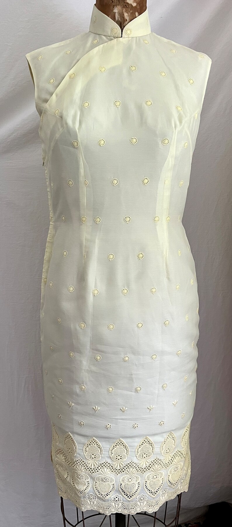 Pode incluir: Um vestido branco sem mangas com gola mandarim e uma bainha de renda festonada. O vestido &eacute; coberto por pequenos c&iacute;rculos bordados brancos.
