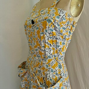 K&ouml;nnte beinhalten: Ein Vintage-Sommerkleid mit einem lebendigen gelben, wei&szlig;en und blauen Blumenmuster. Das Kleid hat einen quadratischen Ausschnitt, breite Tr&auml;ger und zwei Vordertaschen. Schwarze Kn&ouml;pfe zieren das Oberteil und die Taschen und verleihen dem Kleid einen klassischen Touch.