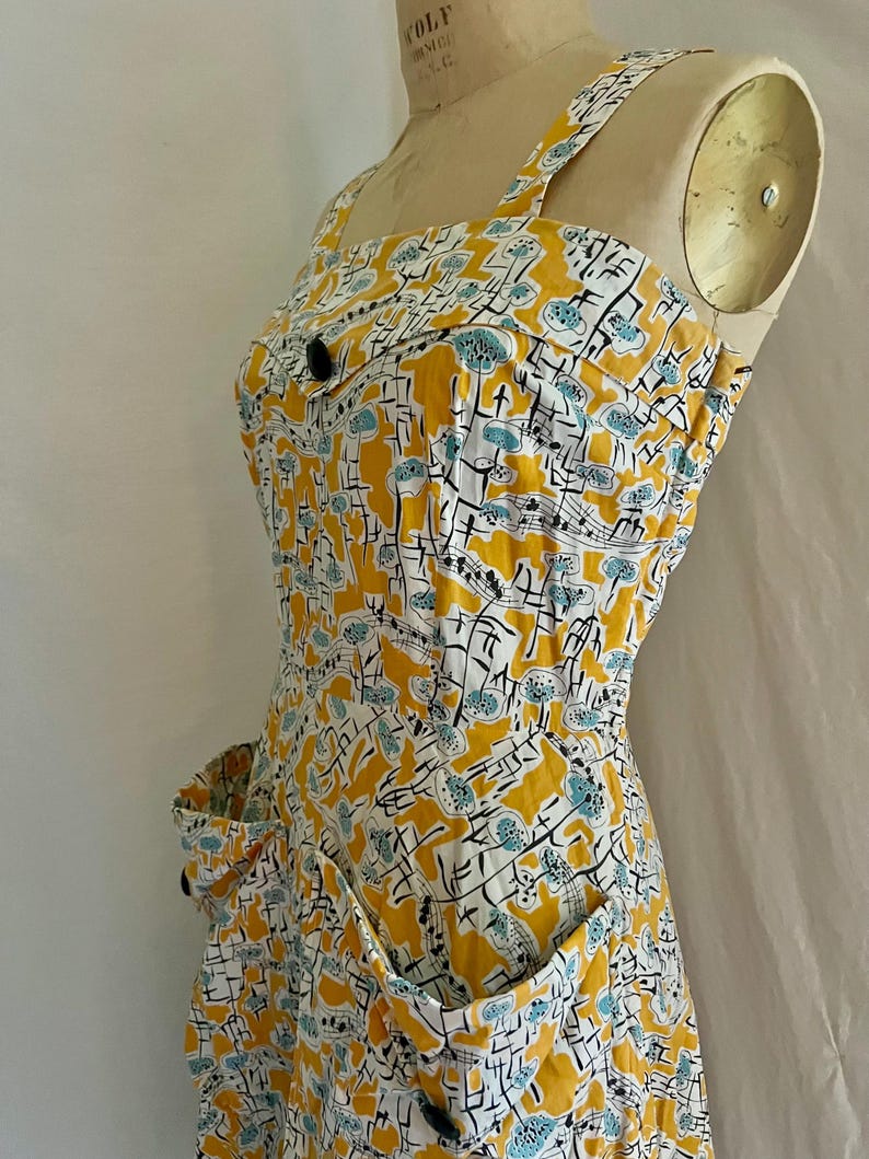 K&ouml;nnte beinhalten: Ein Vintage-Sommerkleid mit einem lebendigen gelben, wei&szlig;en und blauen Blumenmuster. Das Kleid hat einen quadratischen Ausschnitt, breite Tr&auml;ger und zwei Vordertaschen. Schwarze Kn&ouml;pfe zieren das Oberteil und die Taschen und verleihen dem Kleid einen klassischen Touch.