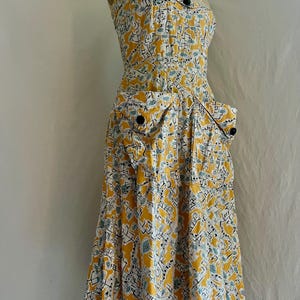 K&ouml;nnte beinhalten: Ein Vintage-Sommerkleid in Gelb, Wei&szlig; und Blau. Das Kleid hat einen einzigartigen Druck, ein eng anliegendes Oberteil und einen ausgestellten Rock. Es hat schwarze Kn&ouml;pfe und zwei Vordertaschen. Das Kleid wird auf einer Schaufensterpuppe pr&auml;sentiert.