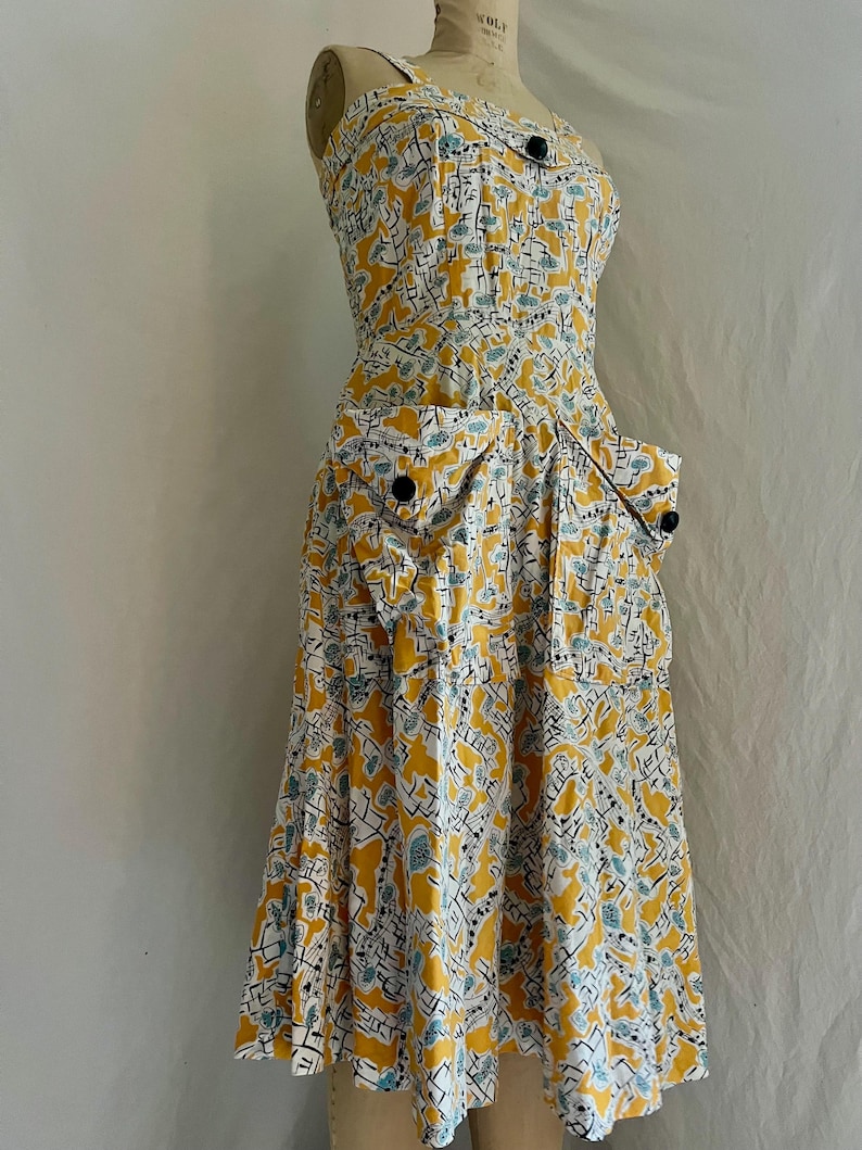 K&ouml;nnte beinhalten: Ein Vintage-Sommerkleid in Gelb, Wei&szlig; und Blau. Das Kleid hat einen einzigartigen Druck, ein eng anliegendes Oberteil und einen ausgestellten Rock. Es hat schwarze Kn&ouml;pfe und zwei Vordertaschen. Das Kleid wird auf einer Schaufensterpuppe pr&auml;sentiert.