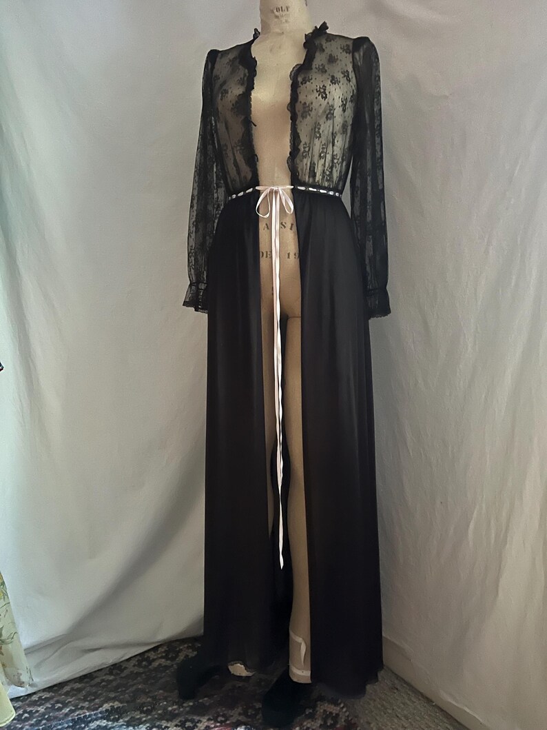 May include: A long, black robe with a sheer, floral lace top and sleeves. The robe has a high collar and a white ribbon tie at the waist. The robe is displayed on a mannequin.