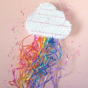 May include: A white cloud-shaped pinata with a rainbow of colourful streamers hanging from the bottom. The pinata is on a pink background.
