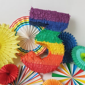 May include: A rainbow-coloured pinata shaped like the number 3. The pinata is surrounded by colourful paper fans.