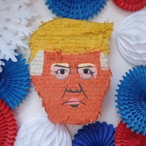 May include: A pinata shaped like a person with a yellow hairpiece and an orange face. The pinata is decorated with blue, red and white paper decorations.