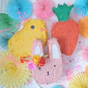 May include: Three colourful pinata decorations: a yellow chick, a pink bunny with a flower crown, and an orange carrot. The pinatas are made of tissue paper and have a fringed texture.