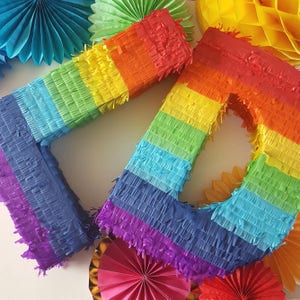 May include: Two rainbow-coloured pinata shaped like the number one. The pinatas are made of tissue paper and have a blue, purple, turquoise, green, yellow, orange, and red colour scheme.