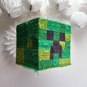 May include: A green and black Minecraft Creeper pinata. The pinata is made of tissue paper and is shaped like a cube. It has a black Creeper face on the front.
