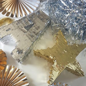 May include: Two gold and silver pinata decorations in the shape of a star and the letter A. The pinatas are made of fringed paper and are hanging from a white background.