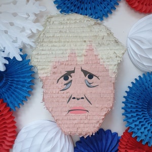 May include: A pink and white pinata shaped like a person with blonde hair. The pinata has a serious expression with a furrowed brow and downturned mouth. The pinata is surrounded by colourful paper decorations.