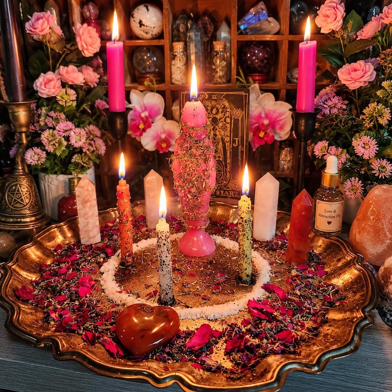 May include: A gold-toned scalloped tray holds a central pink candle covered in dried herbs, surrounded by four smaller lit candles in pink, orange, and yellow. A heart-shaped red stone, white salt, dried flower petals, and crystal points are arranged on the tray.