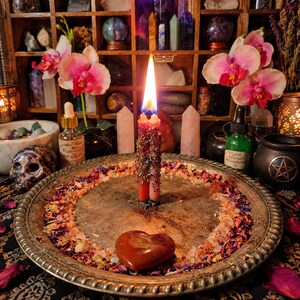 May include: A lit dual-wick candle adorned with dried herbs sits on a round silver tray filled with scattered flower petals and salt crystals. A smooth, heart-shaped red stone rests on the tray, which is surrounded by orchids and crystals.