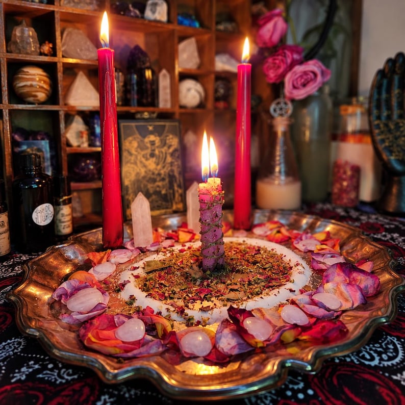 May include: A silver-toned scalloped tray holds three lit candles, including two tall red tapers and one shorter pink candle wrapped in twine. The tray is decorated with scattered dried herbs, flower petals, and small clear quartz crystals.