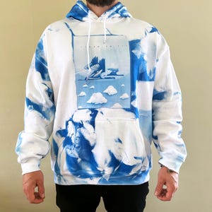 May include: A white hoodie featuring a blue tie-dye cloud pattern throughout. The centre chest displays a rectangular graphic with the text LEARN TO FLY above an illustration of a bird in flight over a landscape with scattered white clouds.