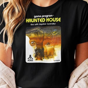 Puede incluir: Una camiseta negra con el gráfico del programa de juego Atari Haunted House. El estampado incluye el texto game program, HAUNTED HOUSE, Use with Joystick Controller, el logotipo de Atari y CX2654, con una ilustración de un murciélago.
