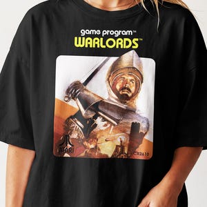 May include: A black t-shirt featuring a graphic print of the Atari Warlords game program. The design displays a knight in armour holding a sword, with the text game program, WARLORDS, the Atari logo, and the model number CX2610 printed on the front.