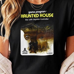 May include: A black t-shirt featuring the Atari Haunted House game program graphic. The print includes the text game program, HAUNTED HOUSE, Use with Joystick Controller, and ATARI CX2654, with an illustration of a bat and a spider.