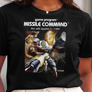 May include: A black t-shirt featuring a graphic print of the Atari Missile Command game box art. The design includes the text game program, MISSILE COMMAND, and Use with Joystick Controller, along with an illustration of a person operating a console.