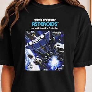 Op de afbeelding: Een zwart t-shirt met een grafische print van het Atari Asteroids spelprogramma. Het ontwerp bevat de tekst game program, ASTEROIDS, Use with Joystick Controller en het Atari-logo met CX2649, met een ruimteschip in de ruimte.