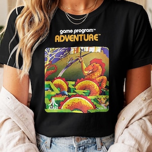 Op de afbeelding: Een zwart t-shirt met de tekst game program ADVENTURE en een kleurrijke grafische illustratie van een draak, een kasteel en een doolhof. Het Atari-logo en de tekst CX2613 staan in de onderste hoeken van het vierkante grafische ontwerp.