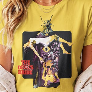 May include: A mustard yellow t-shirt featuring a graphic print of a horned figure holding a person, a large eye, a skull, and other figures. The text "THE DEVIL'S BRIDE" is printed in red on the lower left side of the rectangular black background.
