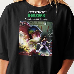 May include: A black t-shirt featuring the Atari Berzerk game program graphic. The print displays the title, the text Use with Joystick Controller, and an illustration of a character in a futuristic setting with a large red robot.