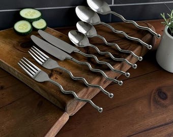 May include: A set of stainless steel flatware featuring a distinct wavy handle design with a small spherical tip. The collection includes two forks, two knives, and three spoons, all arranged in a row on a wooden surface with sliced cucumbers.