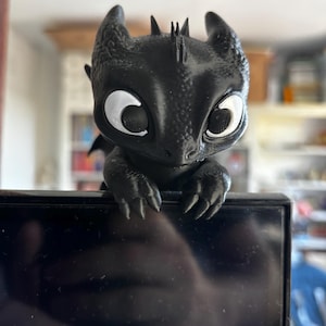 Peut inclure: Une figurine de dragon noire imprimée en 3D avec de grands yeux blancs et des pupilles noires. La créature a une peau texturée, des oreilles pointues et de petites cornes sur la tête. Ses griffes avant reposent sur le bord supérieur d'un écran noir.