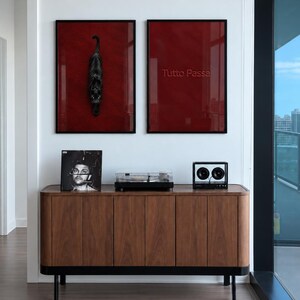 May include: A wooden sideboard with black legs supports a vinyl record, a turntable, and a speaker. Above, two framed prints with deep red backgrounds hang on the wall; one features a black cat, the other the text Tutto Passa.