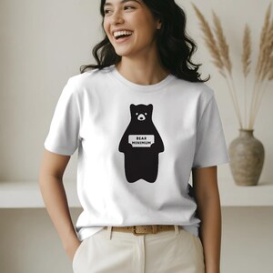 May include: A white short-sleeved t-shirt featuring a black graphic of a bear standing upright. The bear has a white sign on its chest with the black text "BEAR MINIMUM" printed in a clean, sans-serif font. The shirt has a crew neckline.
