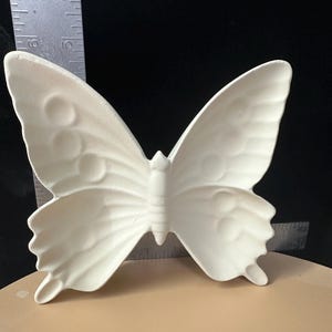 May include: A white, unpainted ceramic butterfly figurine with embossed wing details. A metal ruler is positioned behind the object, showing the butterfly has a height of approximately 6 inches and a width of approximately 6 inches.