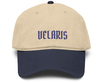May include: A beige baseball cap with a dark navy blue brim and a small matching button on top. The word VELARIS is embroidered in dark navy blue lettering across the front center of the cap, featuring a stylized, gothic-inspired font.