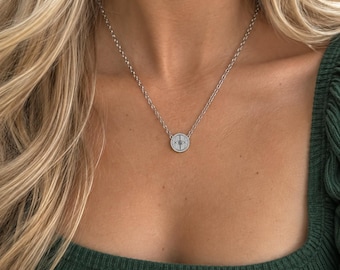 May include: A silver-toned necklace featuring a delicate chain and a small, circular pendant. The pendant has a polished metallic border and a central circular inlay engraved with a geometric pattern consisting of intersecting lines and a small dot.
