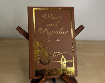 May include: A brown hardcover book titled Pride and Prejudice by Jane Austen, featuring gold foil lettering and a gold illustration of a landscape with a figure. The book is displayed on a small, dark wooden stand with a black ribbon bookmark.