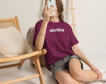 May include: A dark maroon short-sleeved t-shirt featuring the word "witchling" printed in white gothic-style lettering across the chest. The garment is paired with grey denim shorts, creating a casual look with a simple, bold graphic design.
