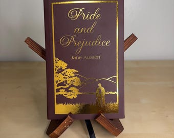 May include: A maroon hardcover book titled Pride and Prejudice by Jane Austen, featuring gold foil lettering and a gold landscape illustration on the front cover, displayed on a small wooden stand with a black ribbon bookmark hanging below.