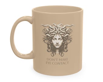 May include: A beige ceramic mug featuring a monochromatic illustration of a figure with snakes for hair, resembling Medusa. Below the illustration, the text reads "DON'T MAKE EYE CONTACT" in a simple, clean, uppercase sans-serif font.