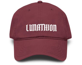 May include: A maroon baseball cap featuring the word LUNATHION embroidered in white capital letters across the front panel. The cap has a structured crown, a curved brim with visible stitching, and a button at the top center of the dome.