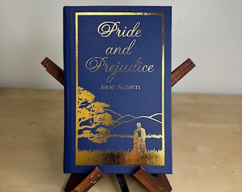 May include: A blue hardcover book titled Pride and Prejudice by Jane Austen, featuring gold foil lettering and a gold landscape illustration of a figure standing near a tree and hills, resting on a small wooden display stand with a black ribbon.