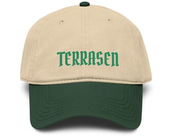 May include: A beige baseball cap with a dark green brim and a dark green button on top. The word TERRASEN is embroidered in dark green capital letters across the front panels of the cap. The cap features stitched eyelets and a curved brim.
