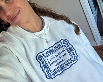 May include: A white crewneck sweatshirt featuring a central rectangular embroidery design in dark blue. The design includes a decorative floral border and the text "I will succeed because I am insane." written in a cursive script font.