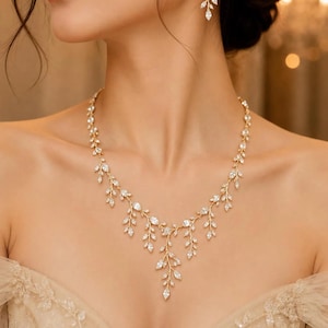Puede incluir: Un conjunto de collar y pendientes colgantes en tono dorado con un delicado diseño de enredadera. La joyería está adornada con numerosos cristales transparentes de talla marquesa dispuestos en un patrón ramificado que imita pequeñas hojas.