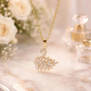 Può includere: Una collana color oro con un ciondolo a forma di cigno. Il ciondolo è incastonato con numerose pietre trasparenti a taglio marquise e rotondo che formano il corpo e il collo del cigno, sospeso a una delicata catena a maglie color oro.