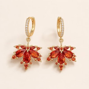 May include: A pair of gold-tone hoop earrings featuring dangling maple leaf charms. Each leaf is encrusted with vibrant orange and red marquise-cut and round-cut crystals. The hoop portion is also detailed with a row of small, clear white crystals.