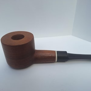 May include: A tobacco pipe featuring a smooth, medium-brown wooden bowl with a cylindrical shape and a wide, flat top. The stem is made of black material, connected to the wooden bowl by a thin, white ring accent at the junction point.