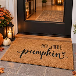 May include: A rectangular tan coir doormat featuring the black printed text "HEY THERE pumpkin" with a small heart symbol. The mat is positioned on a stone porch in front of a black door, surrounded by orange chrysanthemums and pumpkins.
