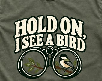 May include: An olive green t-shirt featuring a graphic of black binoculars. Inside the right lens, a small chickadee sits on a brown branch with green leaves. Above the binoculars, the text reads, "HOLD ON, I SEE A BIRD" in bold, cream-colored, distressed lettering.