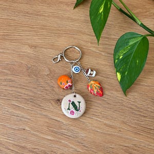 May include: A silver-toned metal keyring featuring a circular beige charm with a green letter N and small pink flowers. Additional charms include an orange sphere with floral patterns, a blue and white evil eye, a coffee cup, and a colourful leaf.