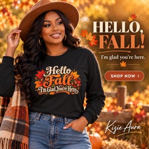 May include: A black long-sleeve shirt featuring a graphic print that says Hello Fall I'm Glad You're Here with colorful autumn leaves and acorns. The shirt is paired with blue denim jeans, a brown wide-brim hat, and a plaid scarf with fringe.