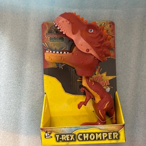 May include: A red and orange T-Rex Chomper dinosaur toy with an oversized head and open mouth, packaged in a yellow and black cardboard display box featuring the text Dinosaur World and T-Rex Chomper with an 8+ ages label on the front.